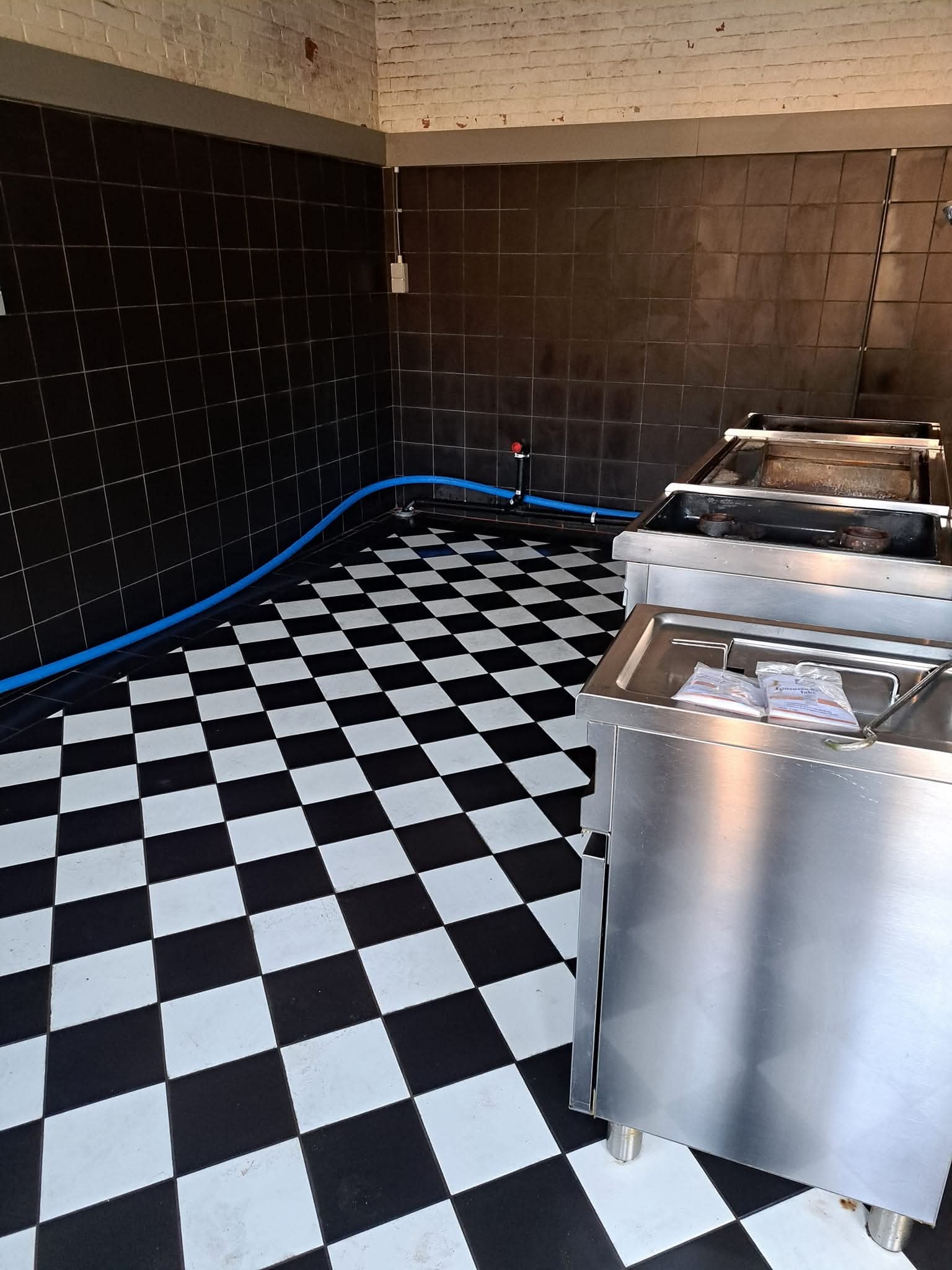 Wnętrze z czarno-białą szachownicą na podłodze i ciemnymi kaflami na ścianach. Widoczne metalowe urządzenia i niebieski wąż. Oświetlenie sufitowe.