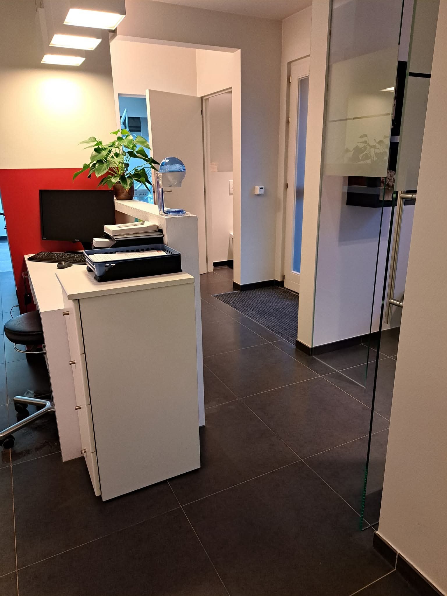 Wnętrze biura z recepcją: białe biurko z komputerem, krzesło obrotowe, roślina doniczkowa. Widok na korytarz z ciemną podłogą i szklanymi drzwiami.