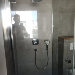 House-Serwis Ireneusz Korzeniewski - Nowoczesna kabina prysznicowa z deszczownicą i szarą armaturą, widoczna przez szklane drzwi. W tle odbicie sylwetki osoby. Minimalistyczny design, szare płytki.