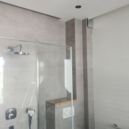 House-Serwis Ireneusz Korzeniewski - Nowoczesna łazienka z kabiną prysznicową typu walk-in, wykończona szarymi płytkami. Widoczne detale: deszczownica, panel sterowania, zabudowa WC z drewnianą półką.