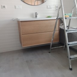 House-Serwis Ireneusz Korzeniewski - Nowoczesna łazienka w trakcie wykańczania: drewniana szafka pod umywalką, okrągłe lustro, szare płytki podłogowe i drabina malarska z gąbką.