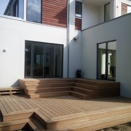 House-Serwis Ireneusz Korzeniewski - Nowoczesny taras z drewna egzotycznego, schody tarasowe prowadzące do domu z białymi ścianami i brązowymi akcentami. Architektura minimalistyczna, jasne drewno.