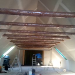 House-Serwis Ireneusz Korzeniewski - Poddasze w trakcie wykańczania: surowe, drewniane belki stropowe, ściany z płyt gips-karton, drabina i narzędzia, pracownik w tle. Wnętrze w jasnych barwach.