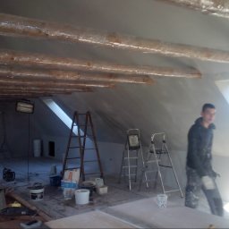 House-Serwis Ireneusz Korzeniewski - Poddasze w trakcie szpachlowania: drewniane belki zabezpieczone folią, drabiny, narzędzia i pracownik w ubraniu roboczym w pobliżu okna dachowego.