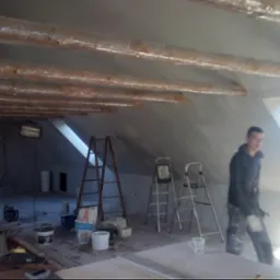 Poddasze w trakcie szpachlowania: drewniane belki zabezpieczone folią, drabiny, narzędzia i pracownik w ubraniu roboczym w pobliżu okna dachowego.