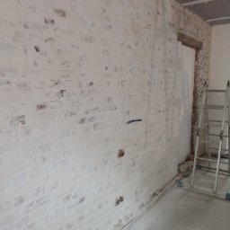 House-Serwis Ireneusz Korzeniewski - Malowana na biało ściana z cegły z widocznymi fragmentami oryginalnej czerwonej cegły, drabina oparta o ścianę, widok na remontowane wnętrze.