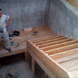 House-Serwis Ireneusz Korzeniewski - Wnętrze w trakcie budowy: drewniana konstrukcja schodów i podestu na tle surowych, szarych ścian. Pracownik z narzędziem w dłoni obserwuje postęp prac.