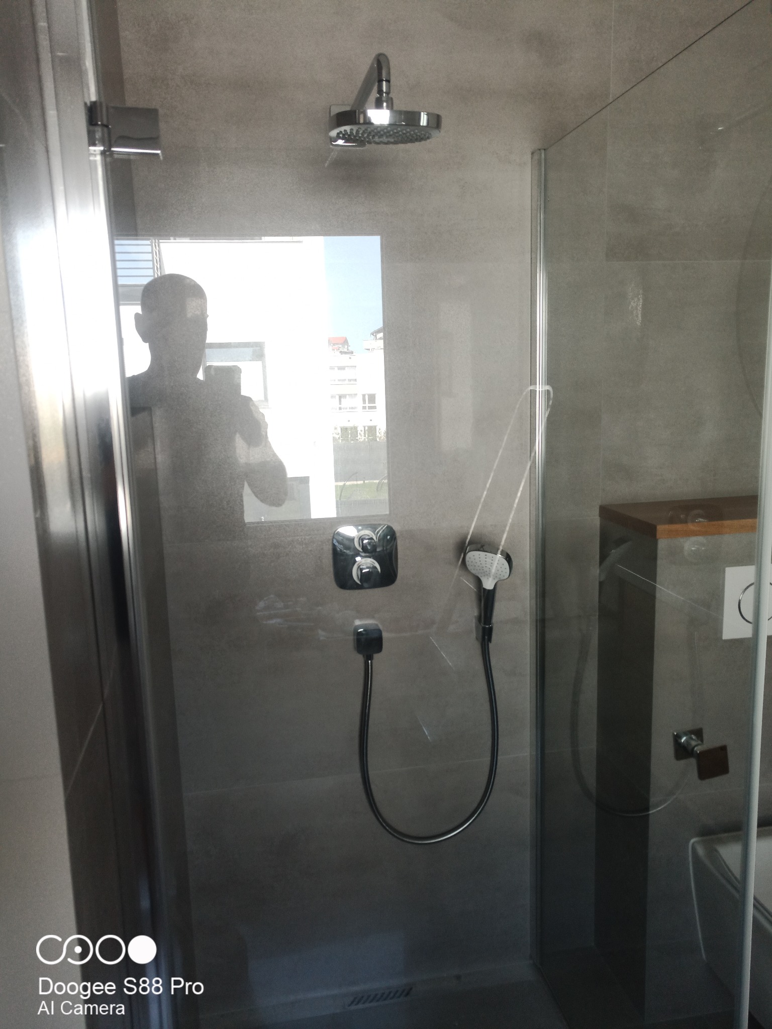 Nowoczesna kabina prysznicowa z deszczownicą i szarą armaturą, widoczna przez szklane drzwi. W tle odbicie sylwetki osoby. Minimalistyczny design, szare płytki.