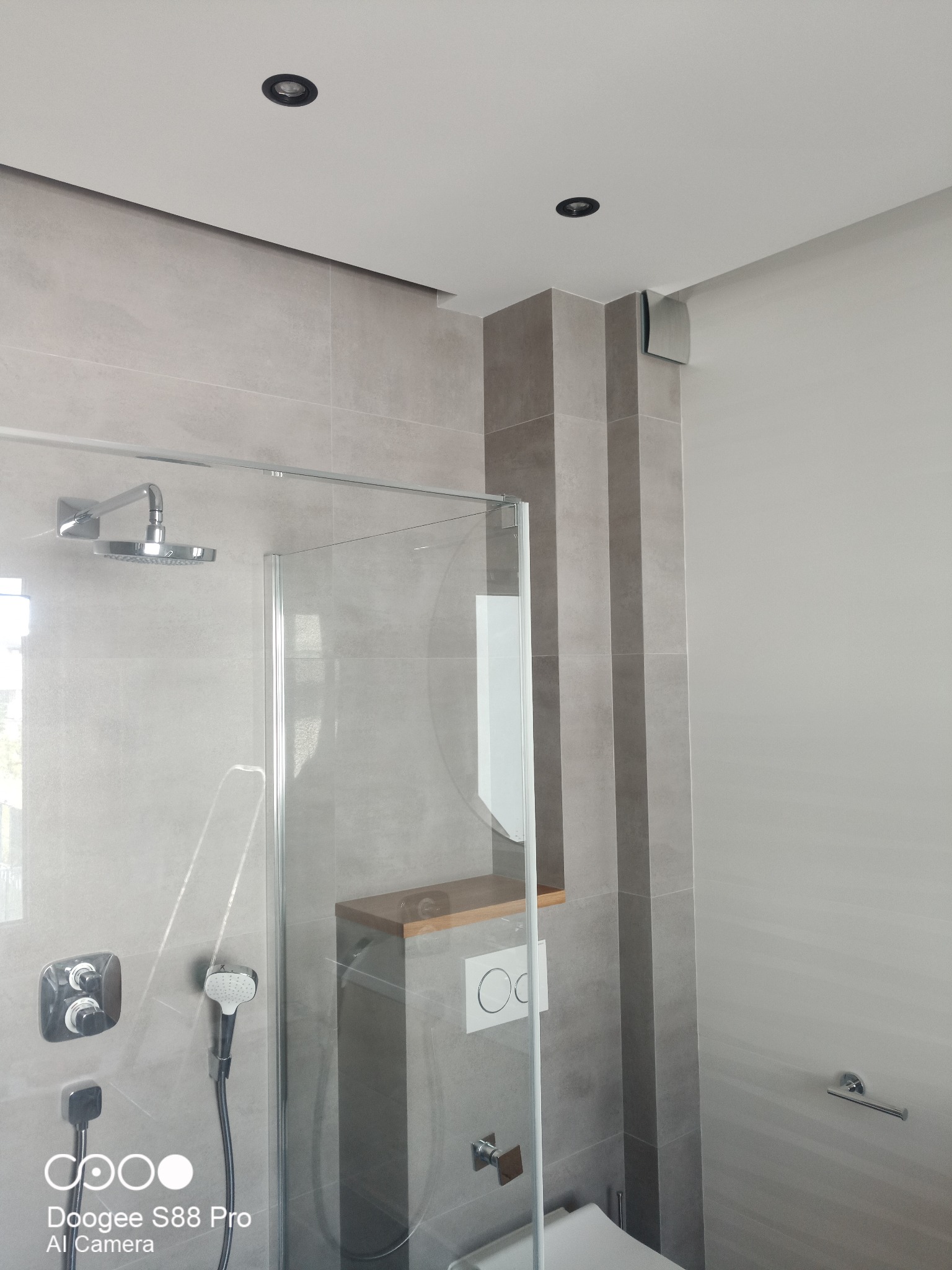 Nowoczesna łazienka z kabiną prysznicową typu walk-in, wykończona szarymi płytkami. Widoczne detale: deszczownica, panel sterowania, zabudowa WC z drewnianą półką.