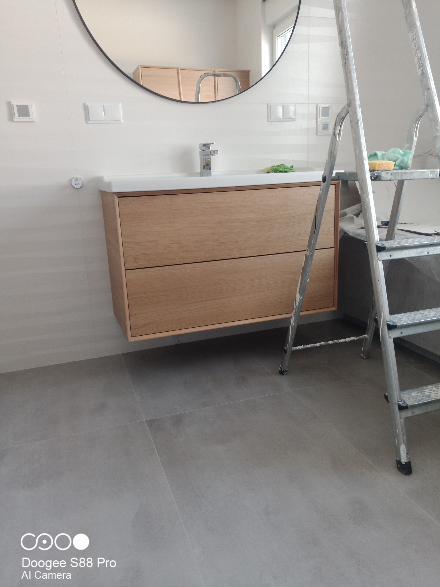 Nowoczesna łazienka w trakcie wykańczania: drewniana szafka pod umywalką, okrągłe lustro, szare płytki podłogowe i drabina malarska z gąbką.