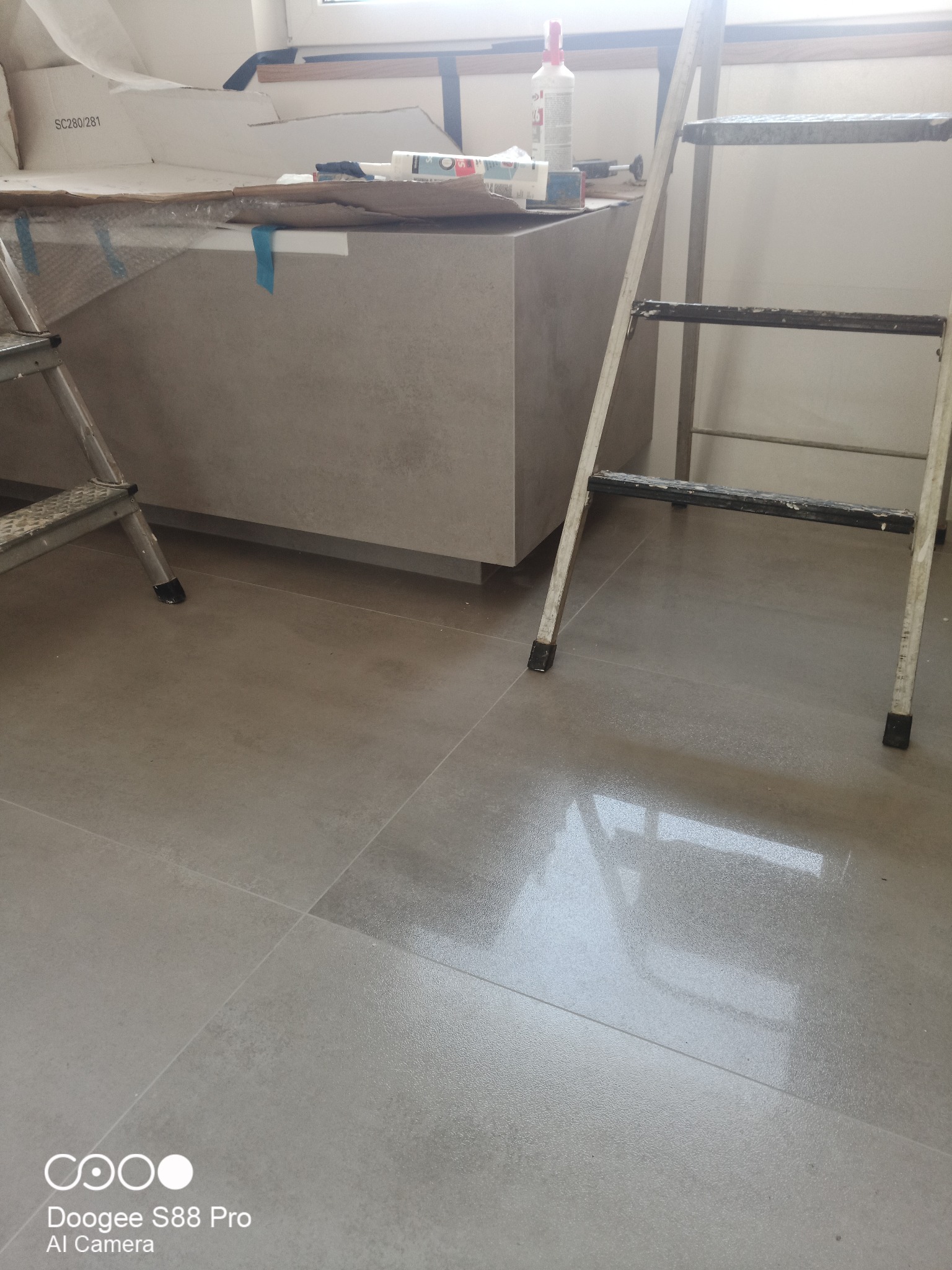 Wykończona, szara zabudowa w łazience, widoczna drabina malarska i błyszczące, szare płytki podłogowe. W tle okno i akcesoria budowlane.