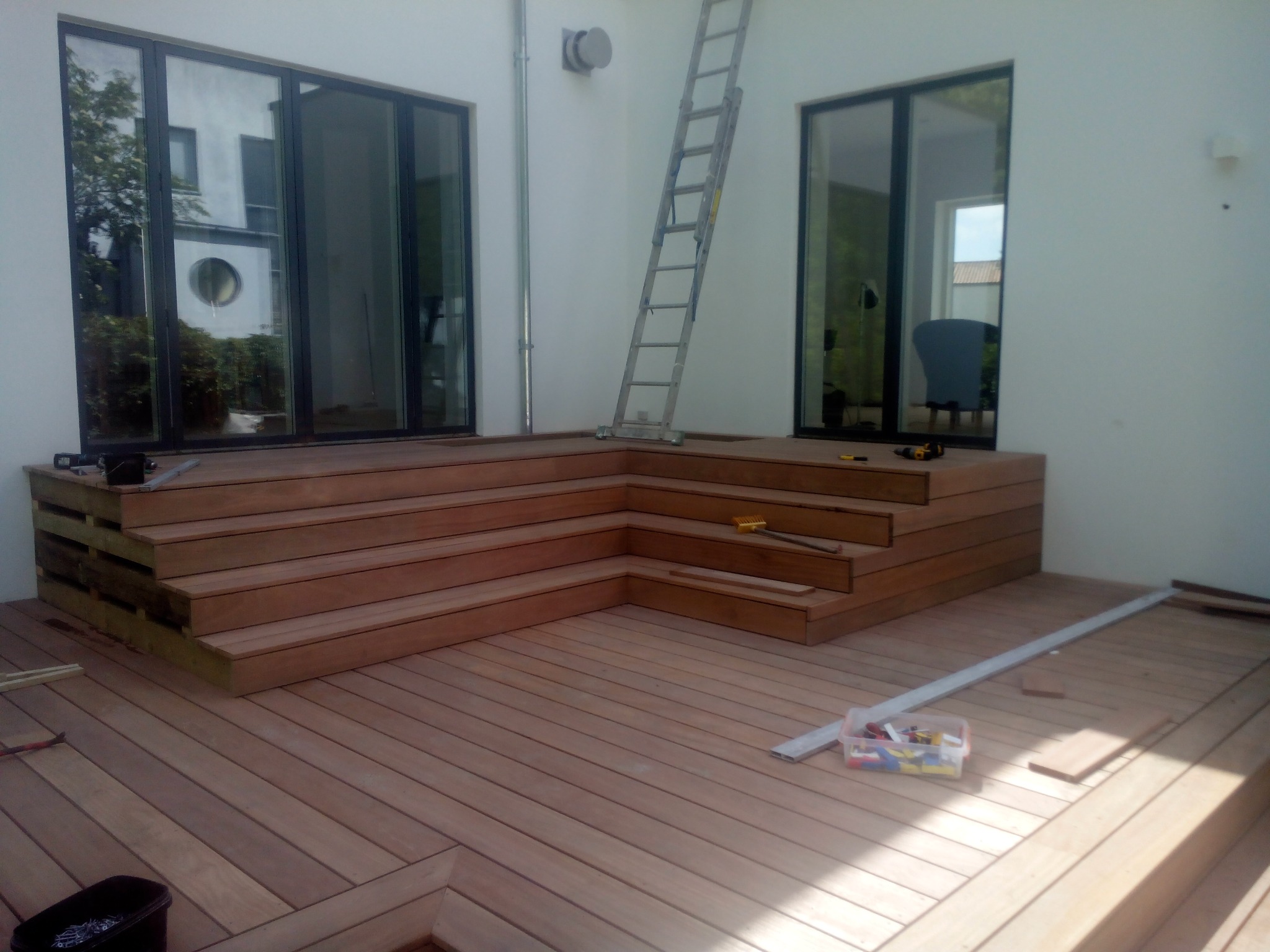 Drewniany taras w trakcie budowy z widocznymi schodami, narzędziami i drabiną opartą o białą ścianę. Przez okna widać zieleń i budynek.