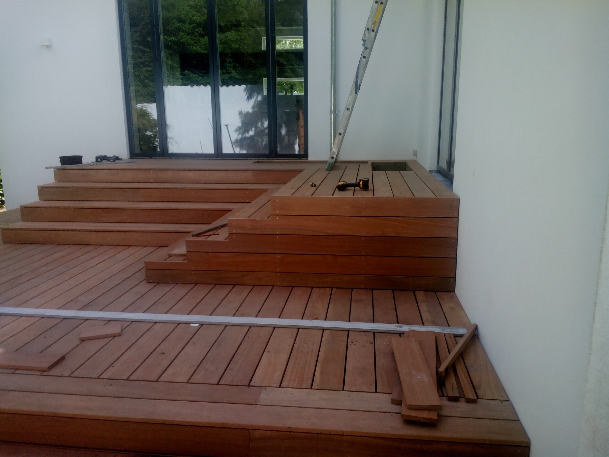 Drewniany taras w trakcie budowy: widoczne deski, schody i narzędzia. Częściowo ukończona konstrukcja z elementami w trakcie montażu, drabina oparta o ścianę.