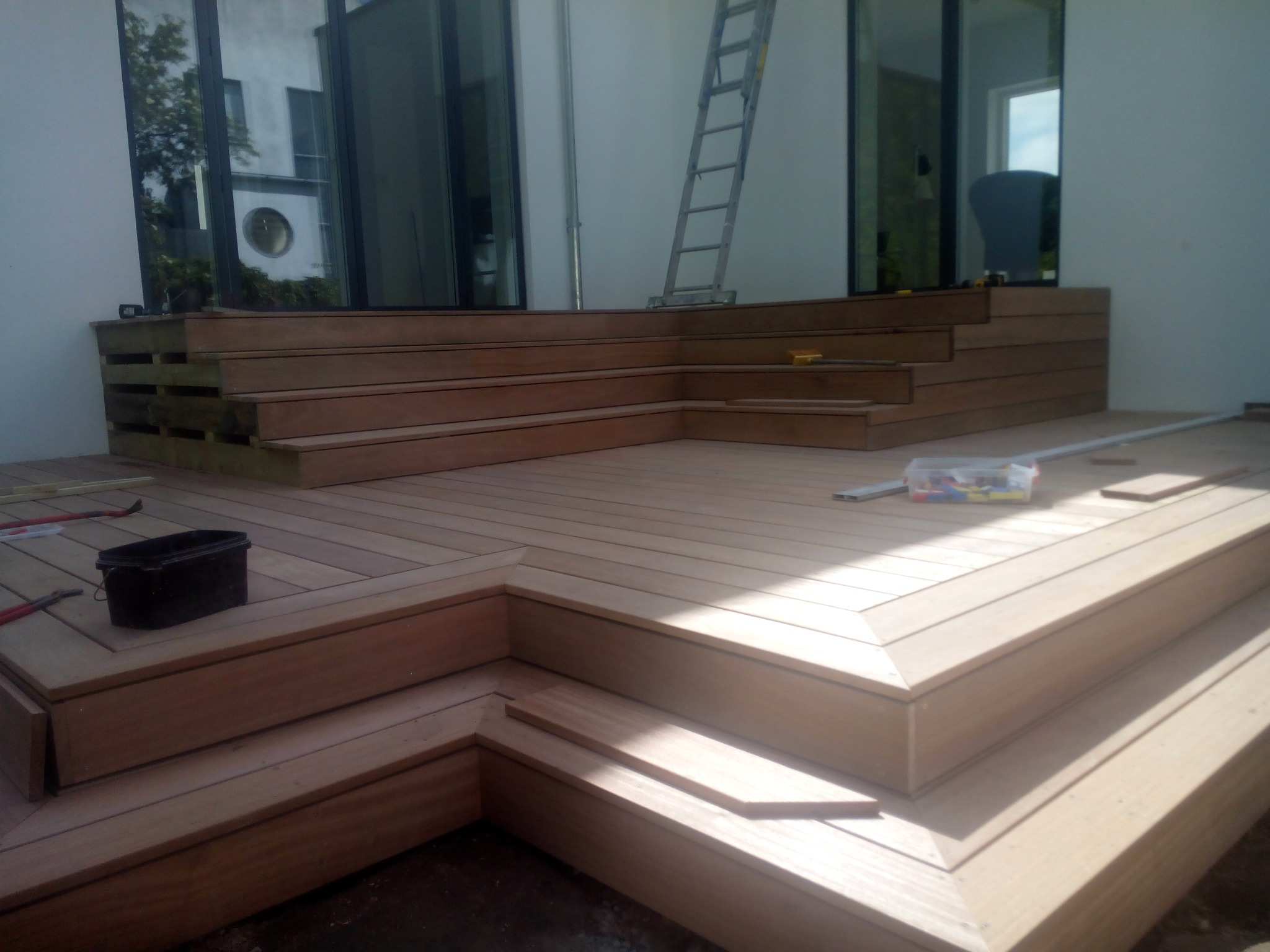 Nowoczesny taras z drewna egzotycznego, z widocznymi schodkami i elementami konstrukcyjnymi. W tle biała ściana budynku i drabina oparta o ścianę.