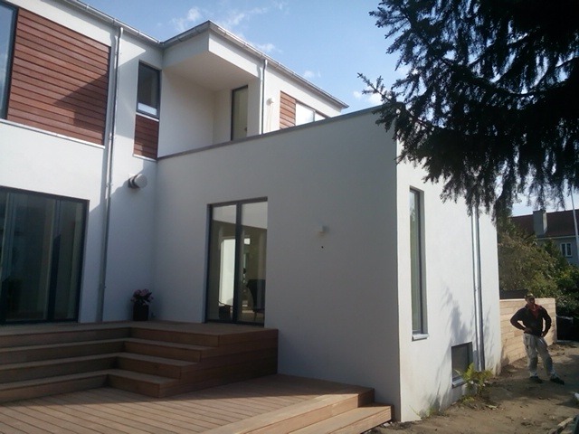 Nowoczesny dom z drewnianym tarasem i szarymi, przesuwnymi oknami. Białe ściany i brązowe elementy elewacji. Widoczna osoba w tle.