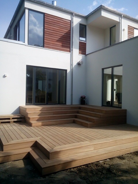 Nowoczesny taras z drewna egzotycznego, schody tarasowe prowadzące do domu z białymi ścianami i brązowymi akcentami. Architektura minimalistyczna, jasne drewno.