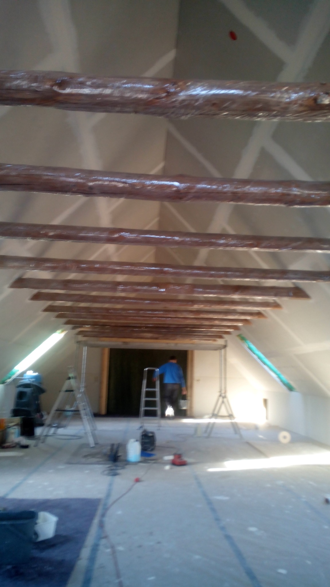 Poddasze w trakcie wykańczania: surowe, drewniane belki stropowe, ściany z płyt gips-karton, drabina i narzędzia, pracownik w tle. Wnętrze w jasnych barwach.