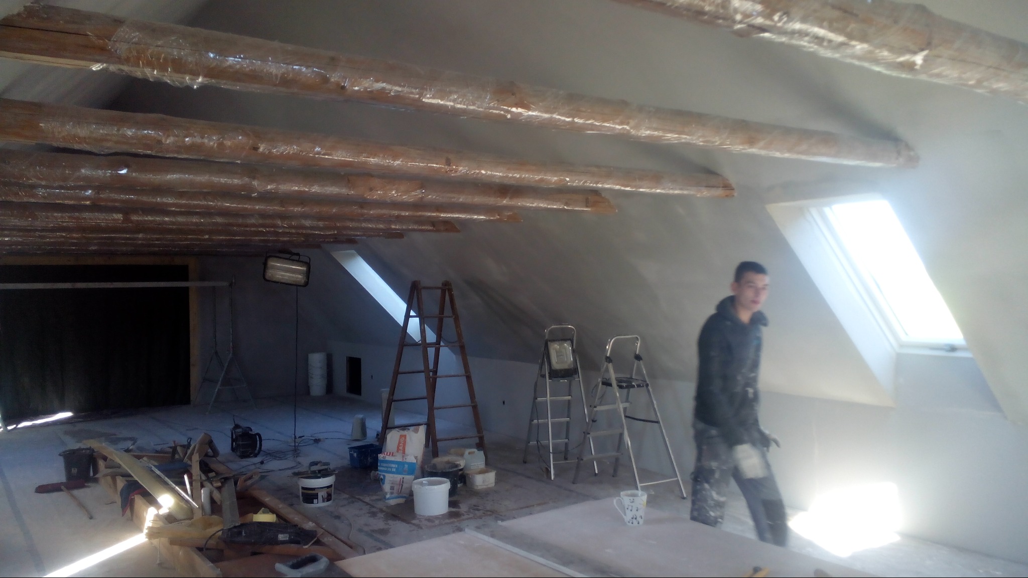 Poddasze w trakcie szpachlowania: drewniane belki zabezpieczone folią, drabiny, narzędzia i pracownik w ubraniu roboczym w pobliżu okna dachowego.
