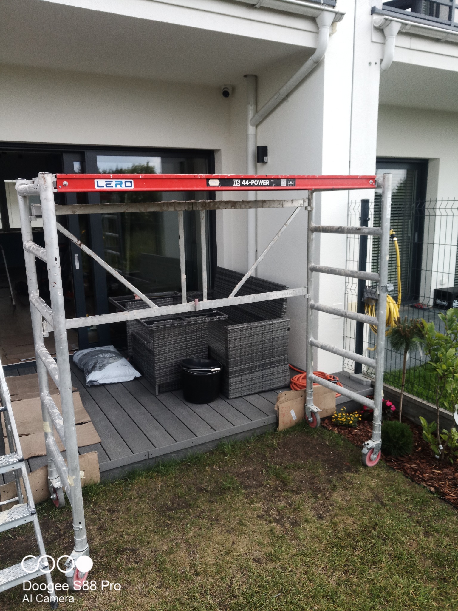 Rozstawiony aluminiowy ruszt na zewnątrz budynku z tarasem, widoczne meble ogrodowe i narzędzia, przygotowanie do prac elewacyjnych lub remontowych na balkonie.