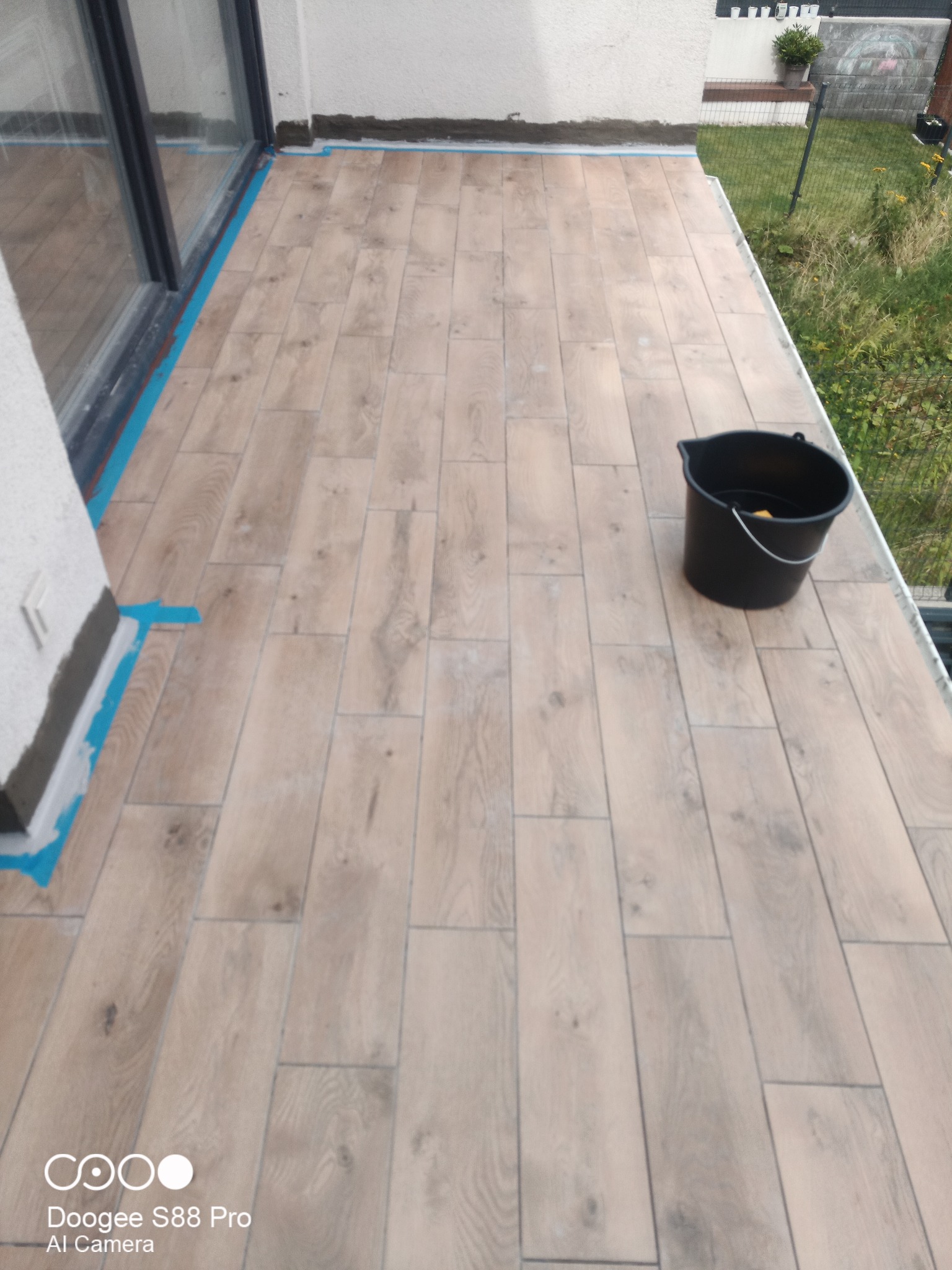 Balkon z wyłożoną jasną płytką imitującą drewno. Widoczne taśmy zabezpieczające przy oknie i ścianie. Czarne wiadro w rogu.