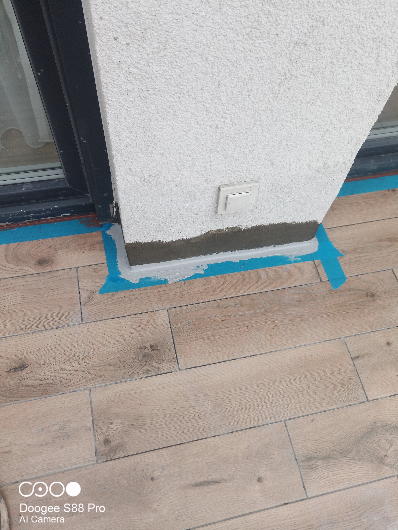 Szczegółowe ujęcie wykończenia balkonu: fuga przy ścianie z gniazdkiem, zabezpieczona niebieską taśmą malarską, na podłodze płytki imitujące drewno.