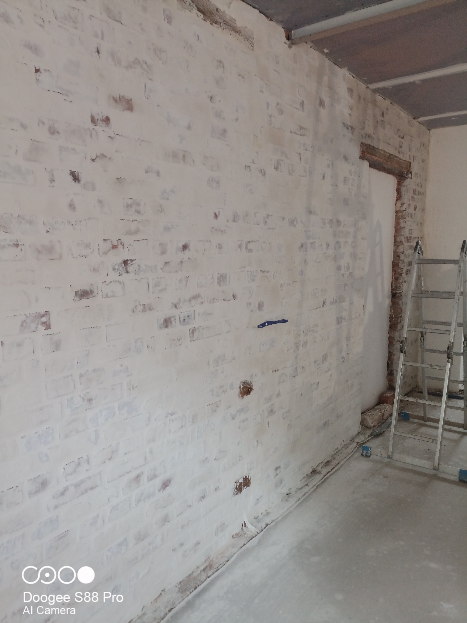 Malowana na biało ściana z cegły z widocznymi fragmentami oryginalnej czerwonej cegły, drabina oparta o ścianę, widok na remontowane wnętrze.