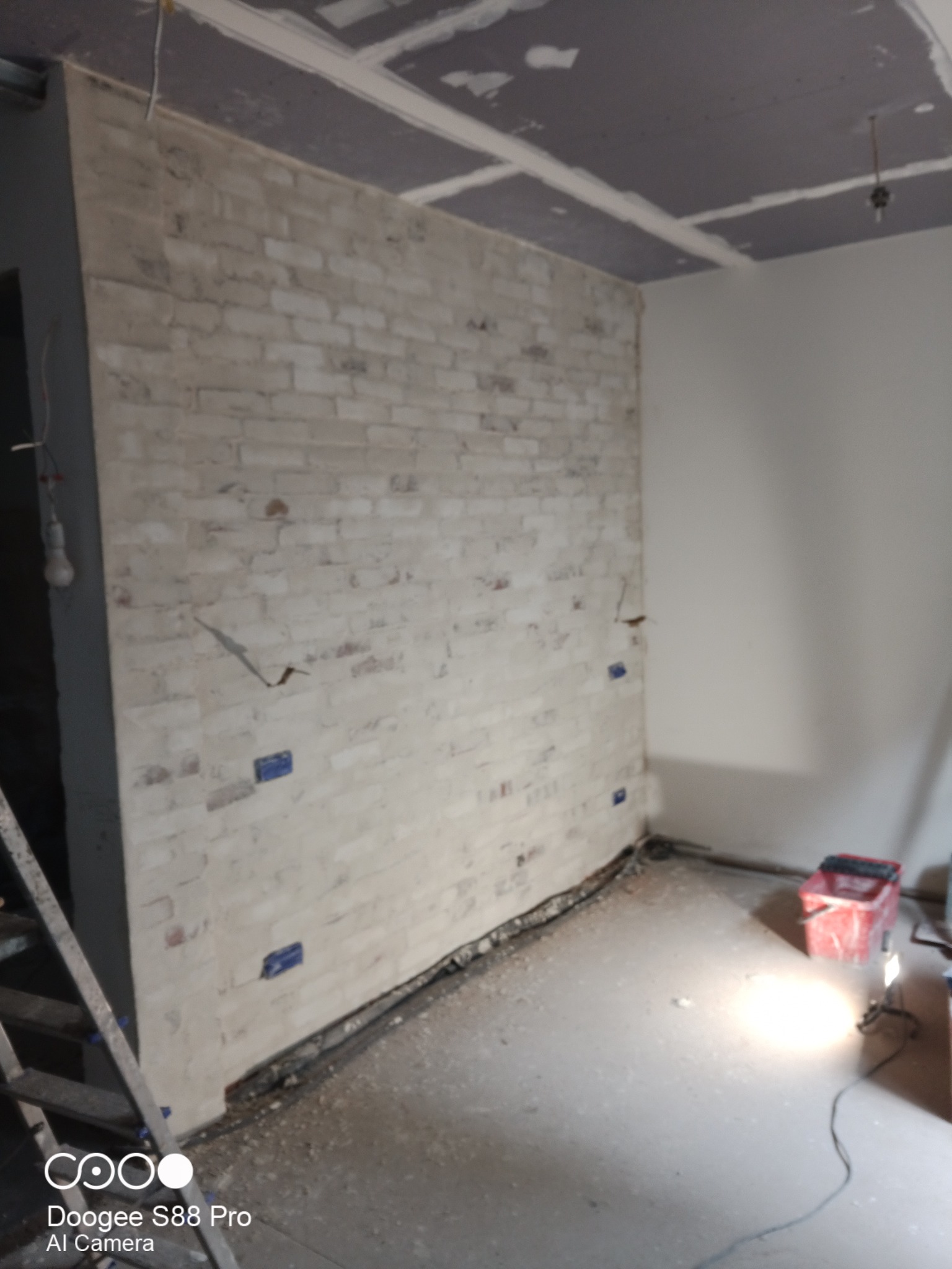 Ściana z cegły dekoracyjnej w trakcie montażu, widoczne niebieskie kliny, sufit z płyt karton-gips, podłoga w stanie surowym, oświetlenie robocze.