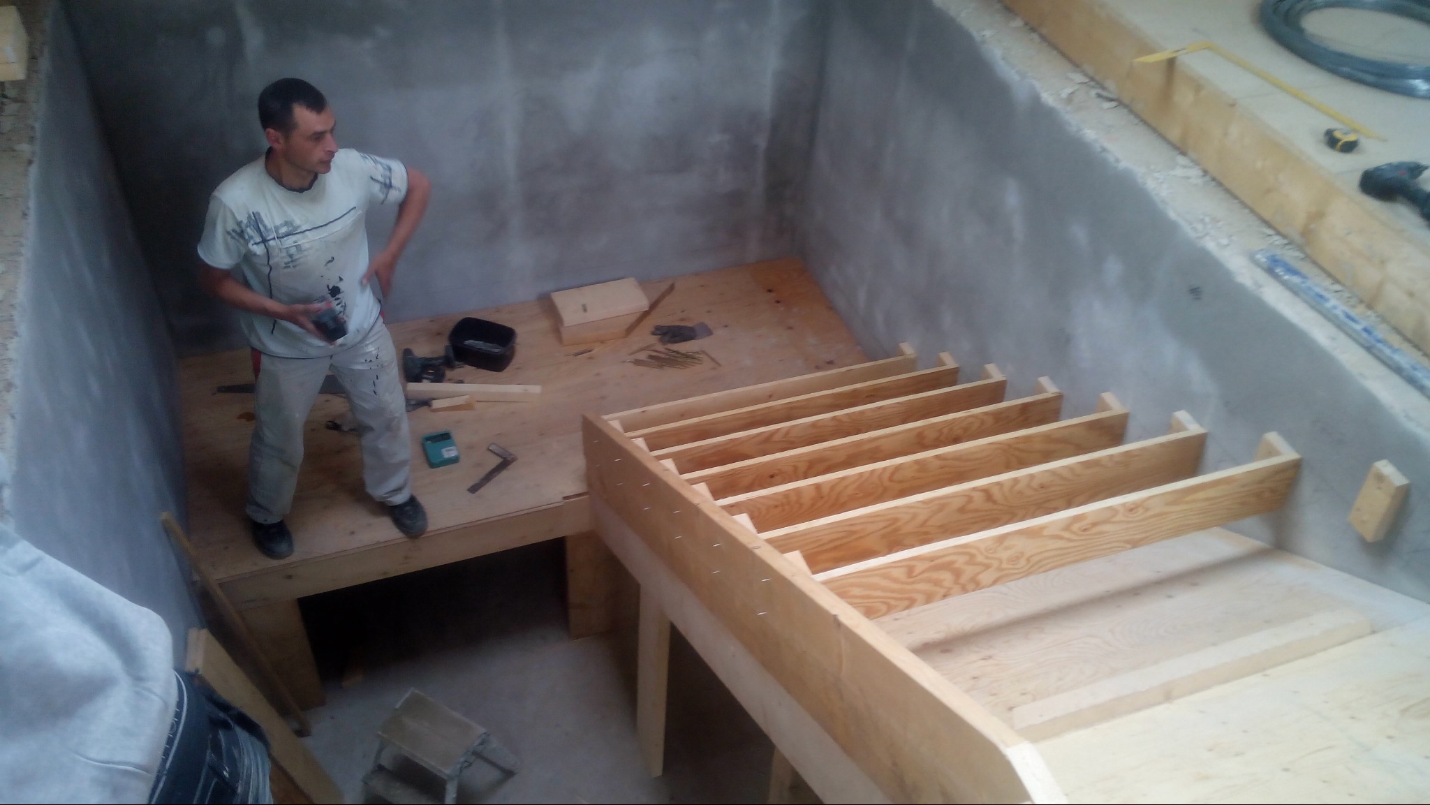 Wnętrze w trakcie budowy: drewniana konstrukcja schodów i podestu na tle surowych, szarych ścian. Pracownik z narzędziem w dłoni obserwuje postęp prac.