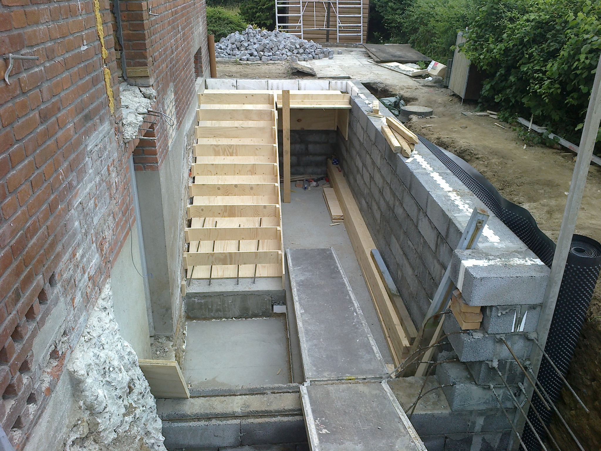 Budowa schodów zewnętrznych z drewnianym szalunkiem i betonowych ścian. Widoczne materiały budowlane i narzędzia. Surowy stan, etap prac ziemnych.