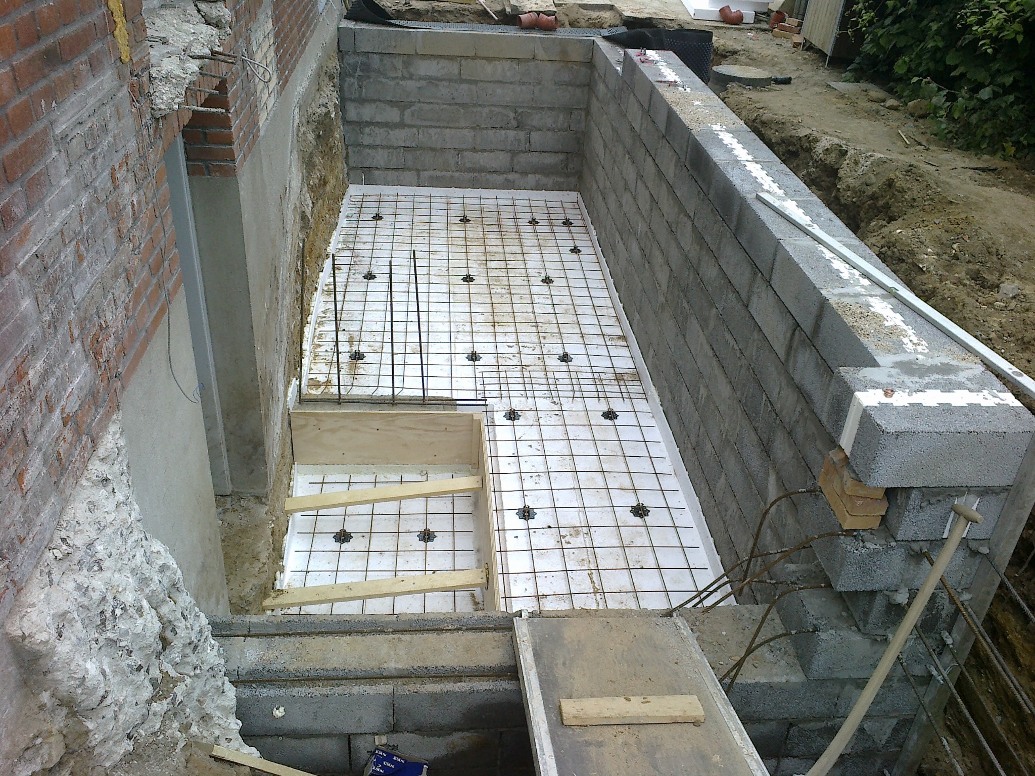 Fundamenty pod budynek w trakcie budowy: ściany z bloczków, izolacja, zbrojenie siatką, częściowo widoczne schody i teren wokół.