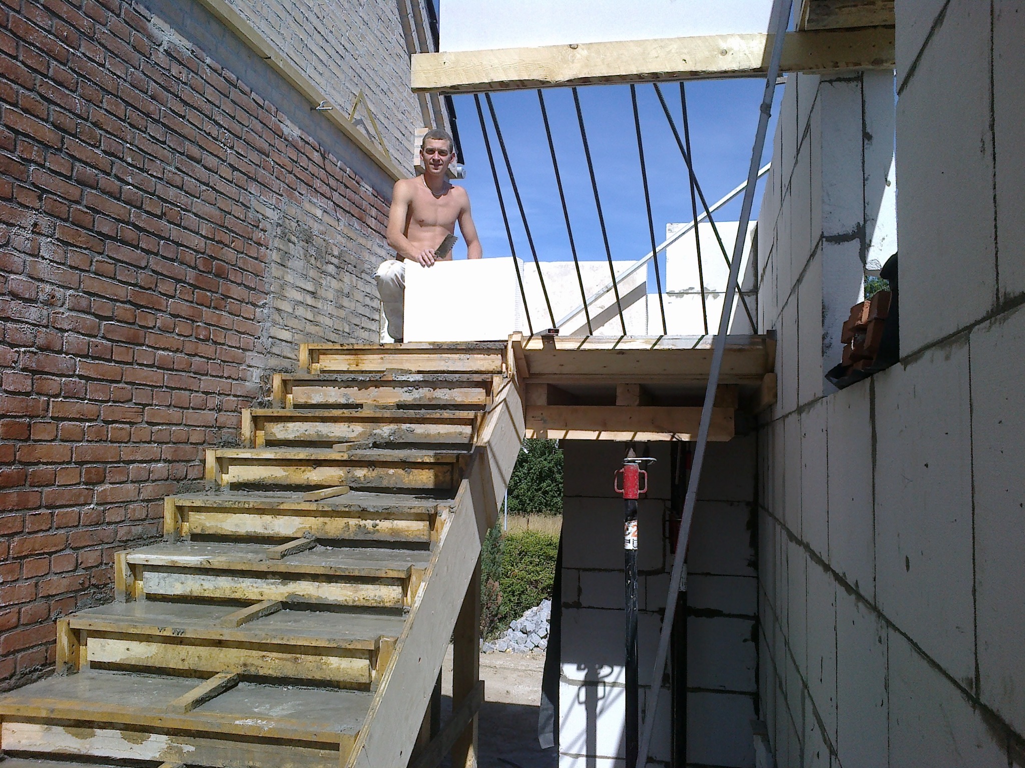 Budowa schodów zewnętrznych z ceglaną ścianą w tle. Mężczyzna w trakcie pracy z bloczkami, widoczne elementy konstrukcyjne i podpora.