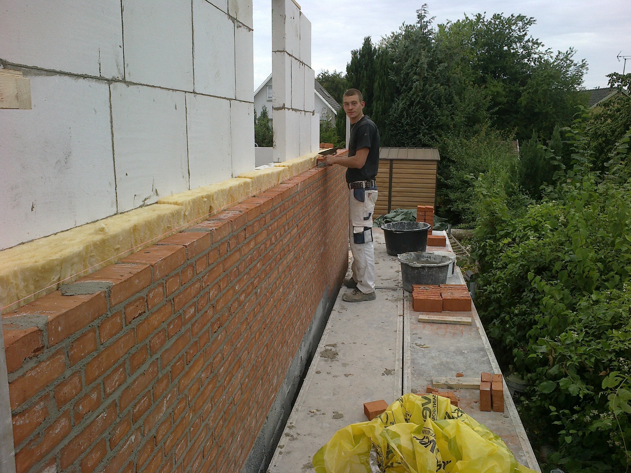 Mężczyzna muruje ścianę z czerwonej cegły, z izolacją, na tle zieleni. Widoczne narzędzia i materiały budowlane. Praca w toku, budowa domu.
