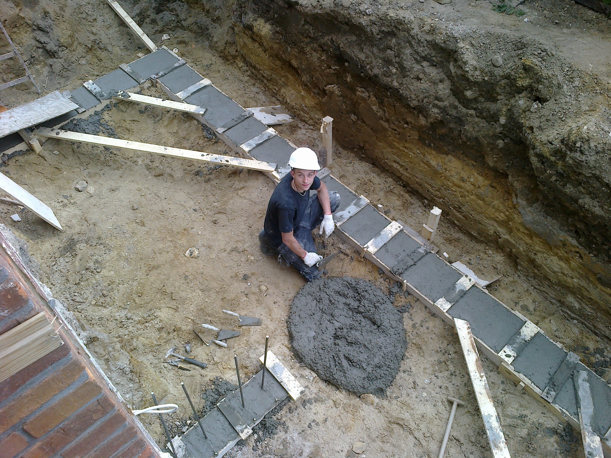 Pracownik w kasku przy wylewaniu fundamentów pod budynek. Widoczne szalunki, beton i narzędzia. Ujęcie z góry, ukazujące etap budowy.