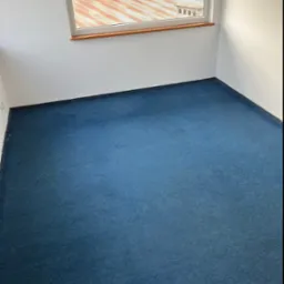 Pokój z niebieską wykładziną dywanową, widok na zewnątrz przez okno. W tle widać budynki i zieleń. Wykładzina czysta, bez plam. Całość w jasnych, naturalnych barwach.