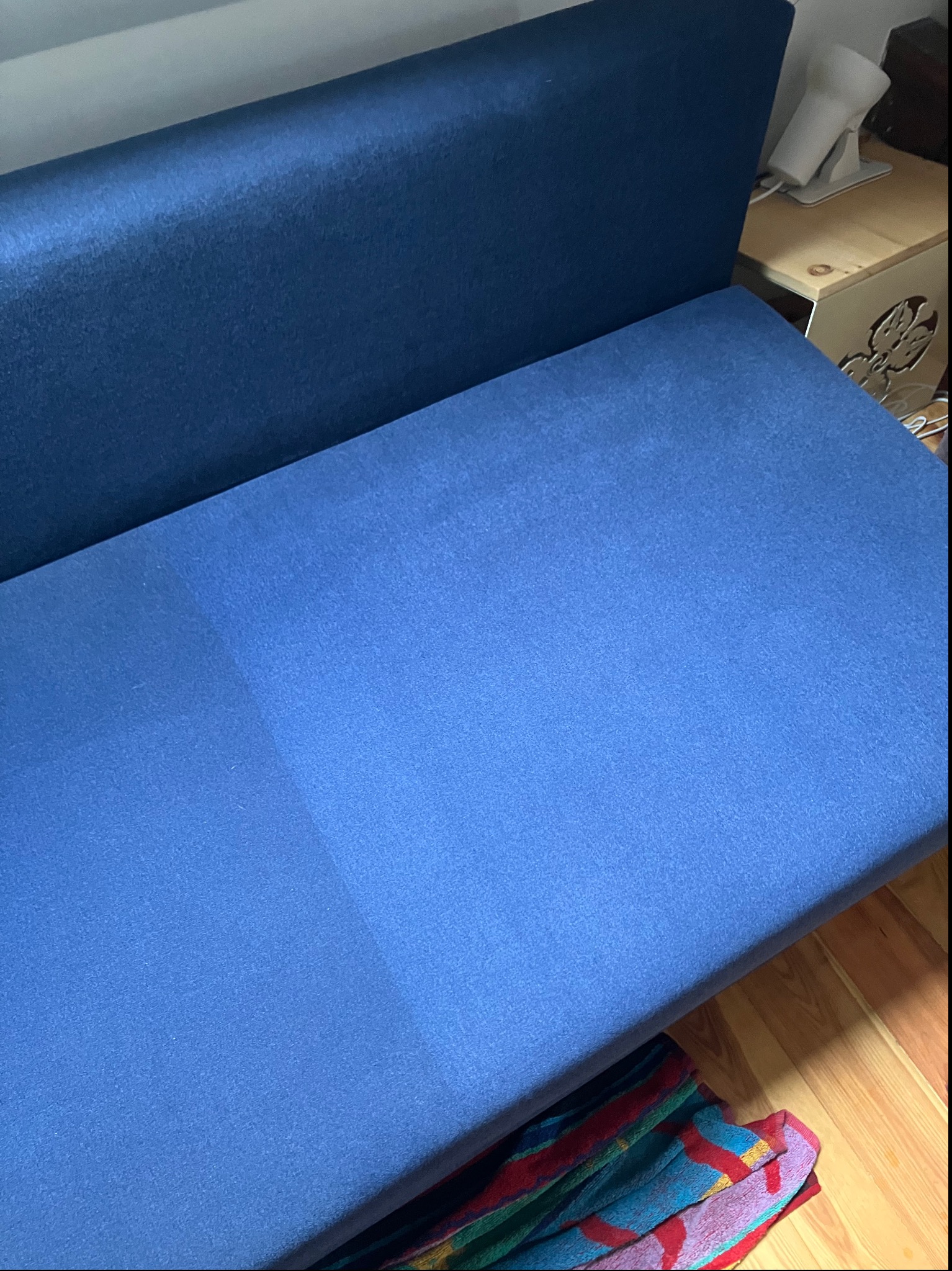 Niebieska kanapa z wyraźną różnicą w odcieniu po czyszczeniu, widoczna jaśniejsza, odświeżona część tapicerki. Ujęcie z góry, w tle fragment podłogi i stolika.