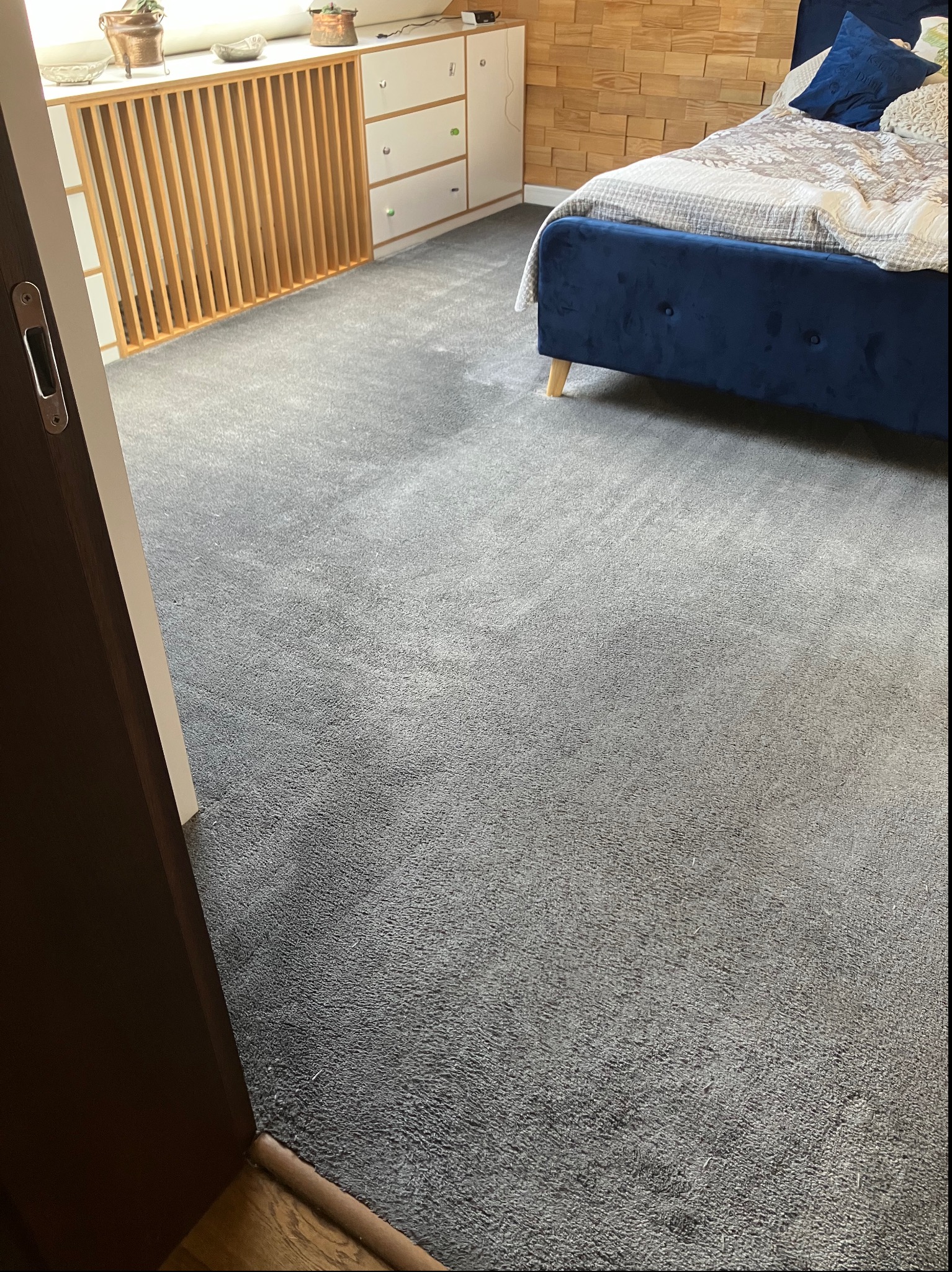 Szary dywan w sypialni z granatowym łóżkiem, widoczne jaśniejsze plamy. Ujęcie z niskiej perspektywy, w tle drewniana ściana i biała komoda.