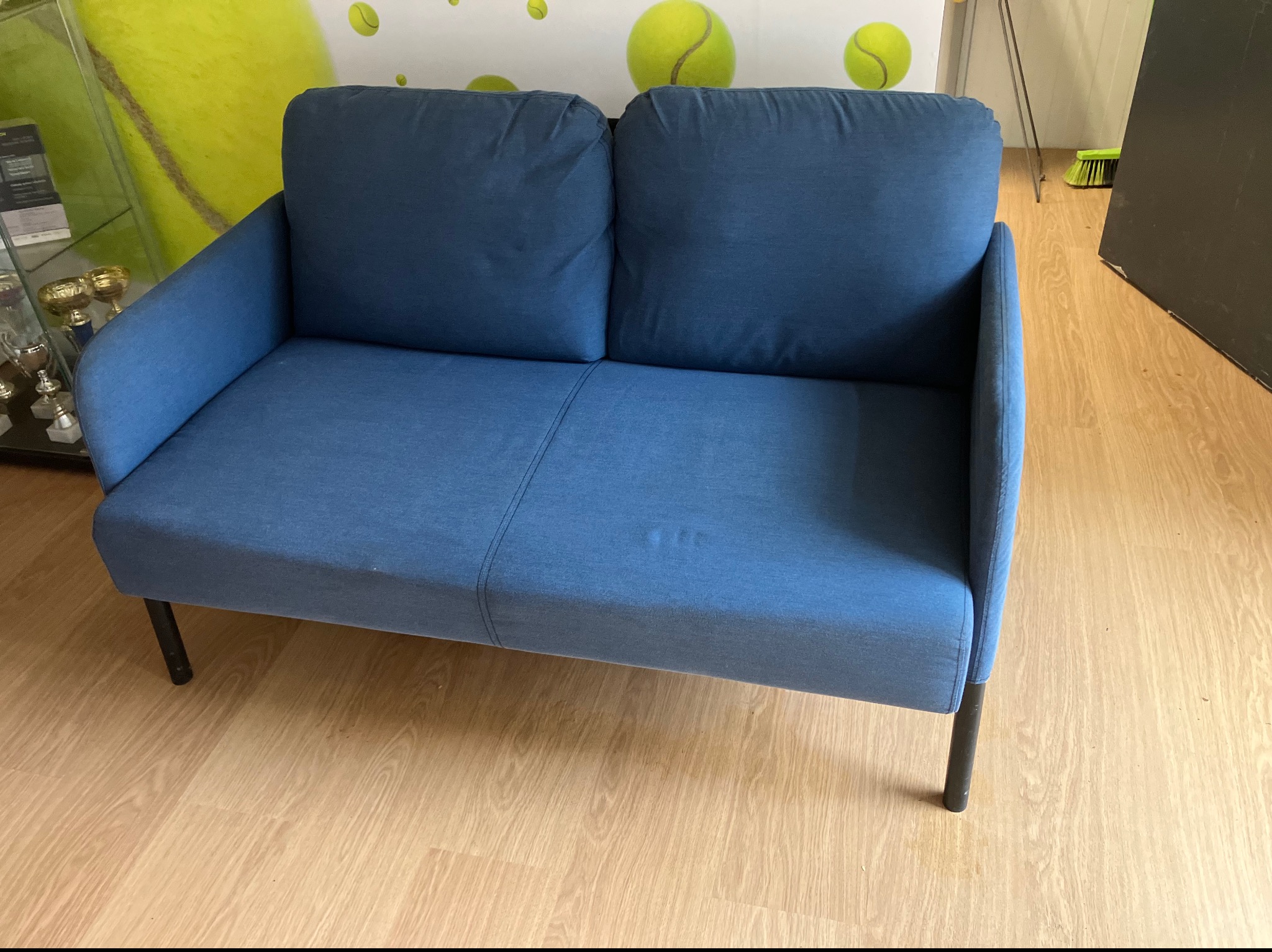 Niebieska sofa dwuosobowa z plamami na siedzisku, stojąca na drewnianej podłodze. W tle ściana z motywem piłek tenisowych i witryna z trofeami.