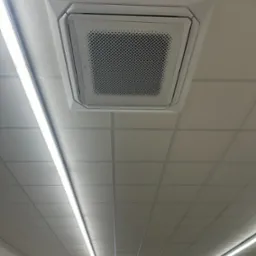 Biała, sufitowa klimatyzacja kasetonowa Fujitsu, wkomponowana w rastrowy sufit podwieszany. Widoczne panele sufitowe i lampa liniowa LED. Ujęcie z dołu.