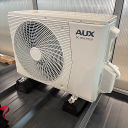 Wentotermika-klimatyzacja Warszawa - Biała jednostka zewnętrzna klimatyzacji AUX DC Inverter zamontowana na czarnych podkładkach antywibracyjnych na metalowym dachu. Widoczne śruby mocujące i przewody.