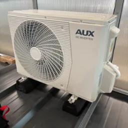 Biała jednostka zewnętrzna klimatyzacji AUX DC Inverter zamontowana na czarnych podkładkach antywibracyjnych na metalowym dachu. Widoczne śruby mocujące i przewody.