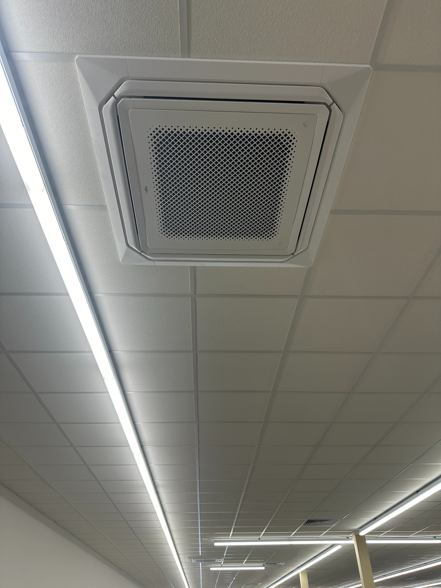 Biała, sufitowa klimatyzacja kasetonowa Fujitsu, wkomponowana w rastrowy sufit podwieszany. Widoczne panele sufitowe i lampa liniowa LED. Ujęcie z dołu.