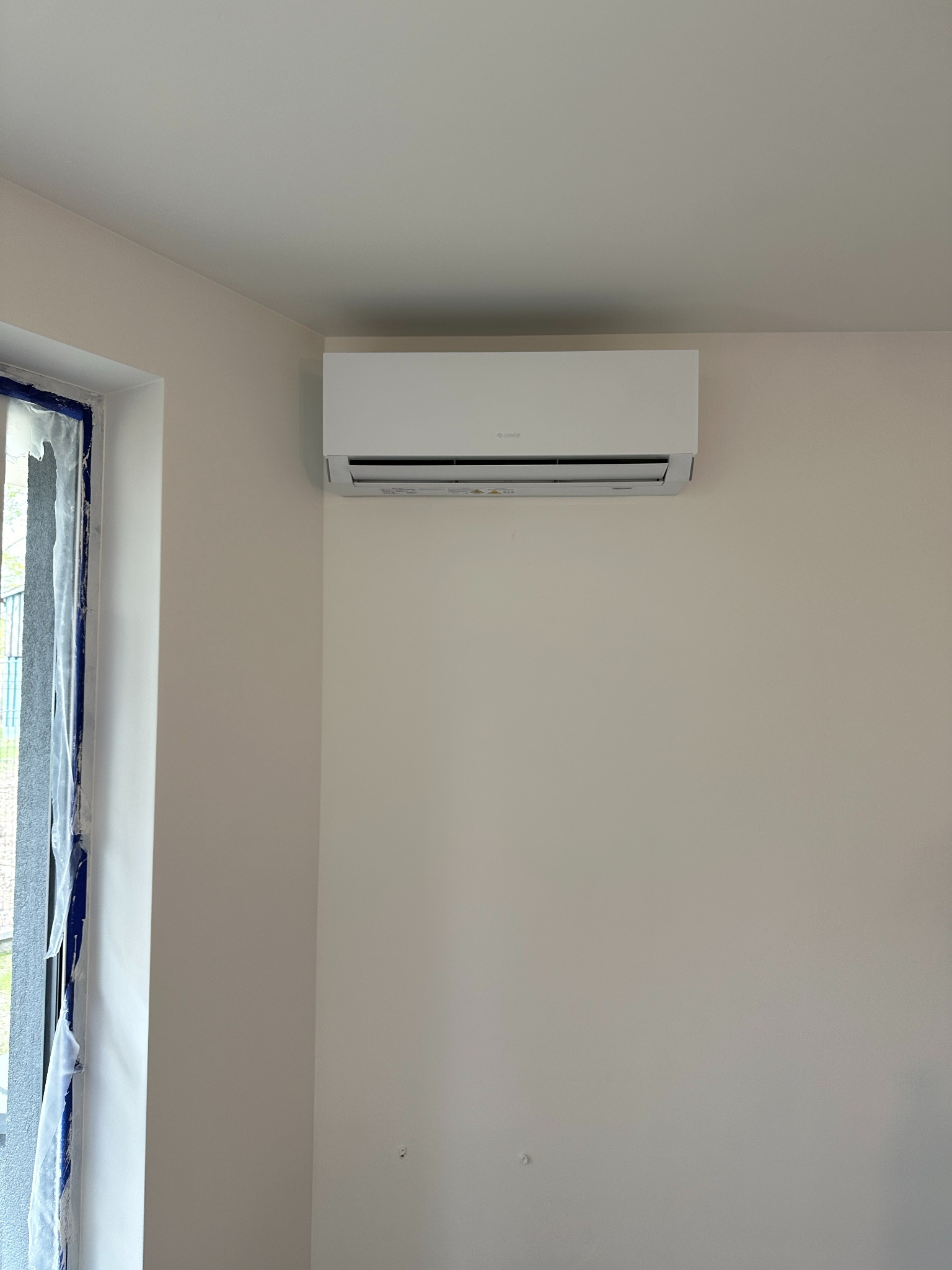 Biała klimatyzacja marki Gree zamontowana na jasnej ścianie w nowym pomieszczeniu z oknem zabezpieczonym folią malarską. Minimalistyczny design.