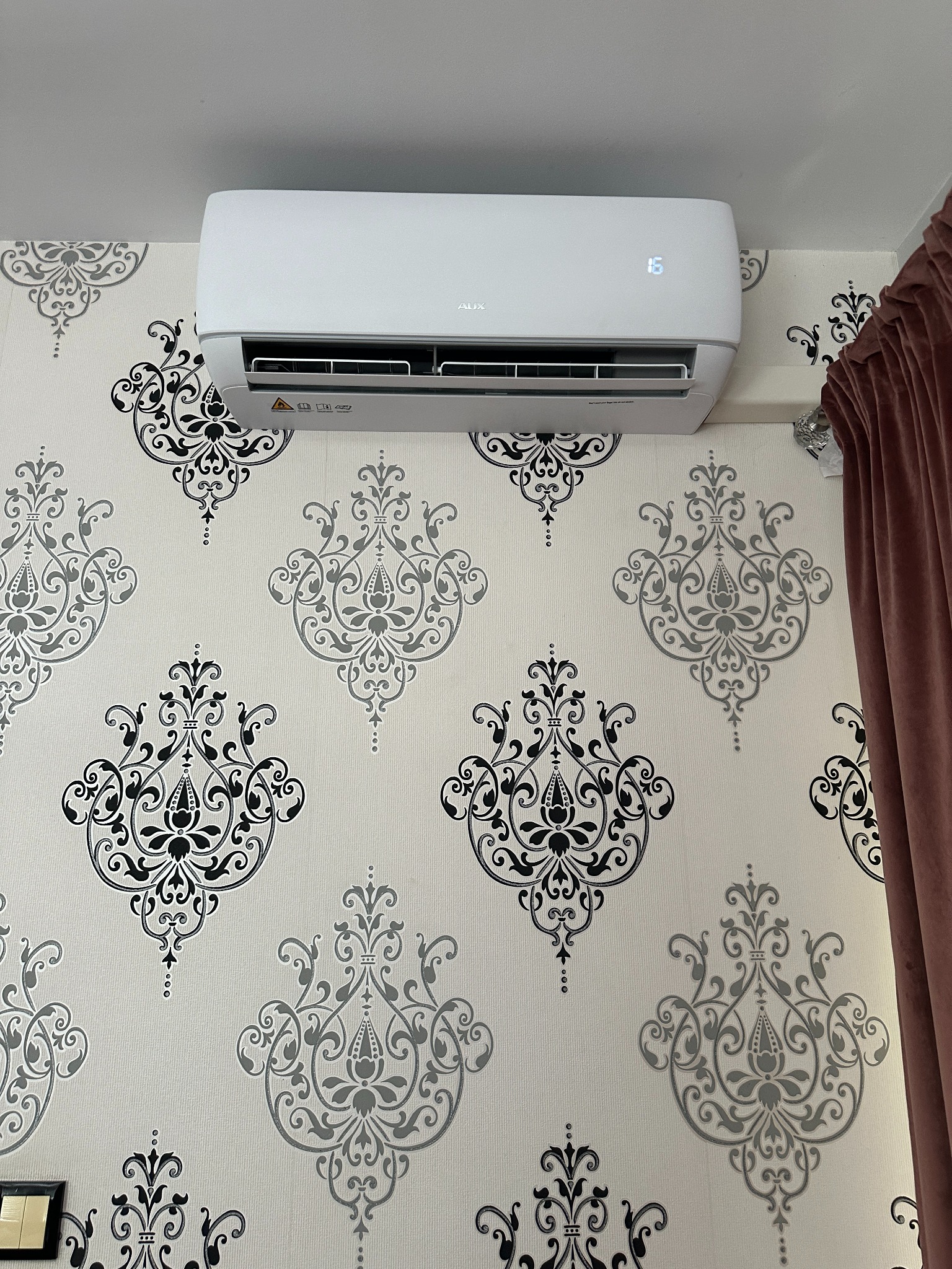 Biała klimatyzacja AUX na ścianie w ozdobne, szaro-czarne ornamenty. Widoczny fragment zasłony po prawej. Na wyświetlaczu urządzenia widoczna temperatura 16 stopni.