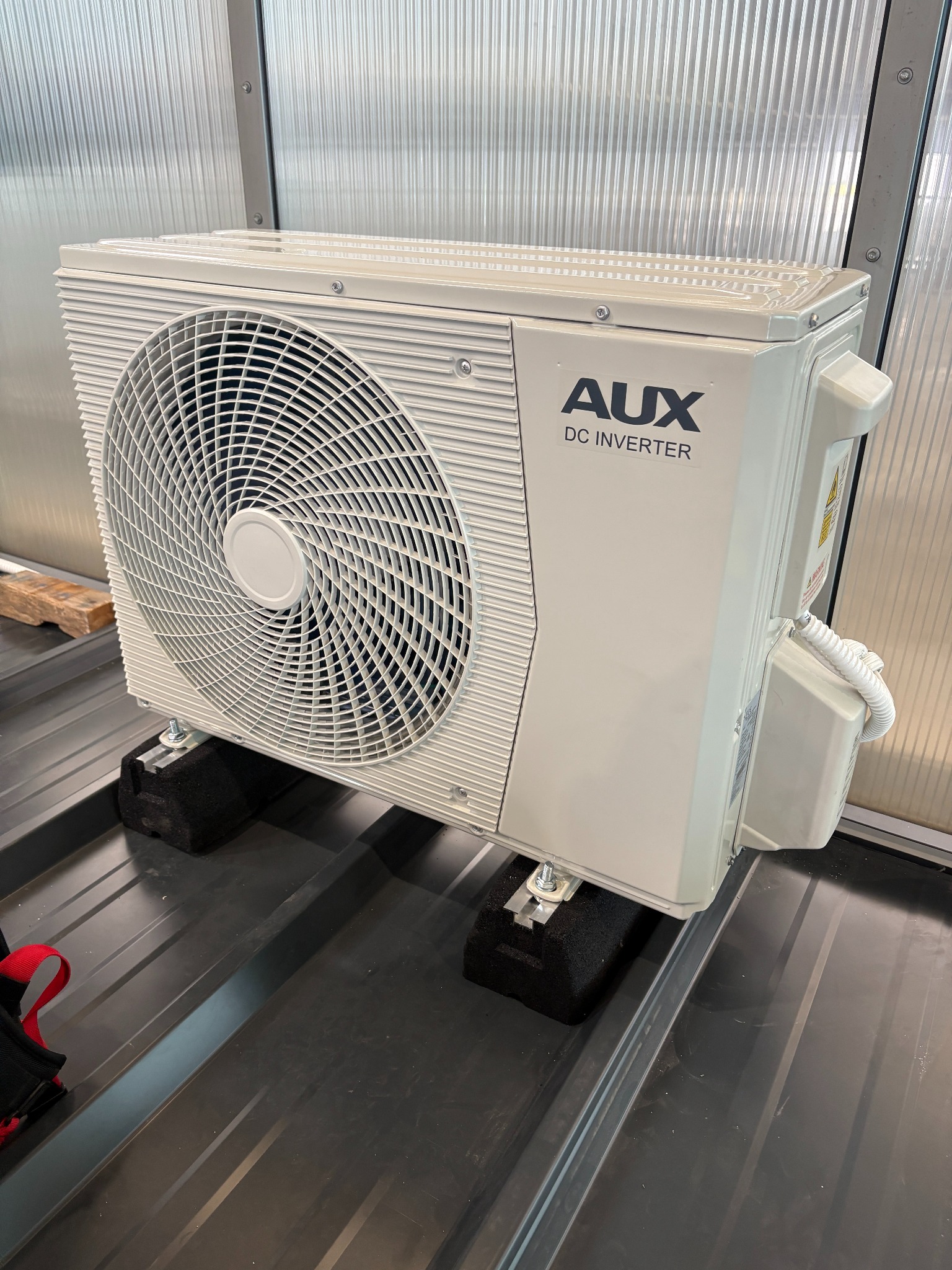 Biała jednostka zewnętrzna klimatyzacji AUX DC Inverter zamontowana na czarnych podkładkach antywibracyjnych na metalowym dachu. Widoczne śruby mocujące i przewody.