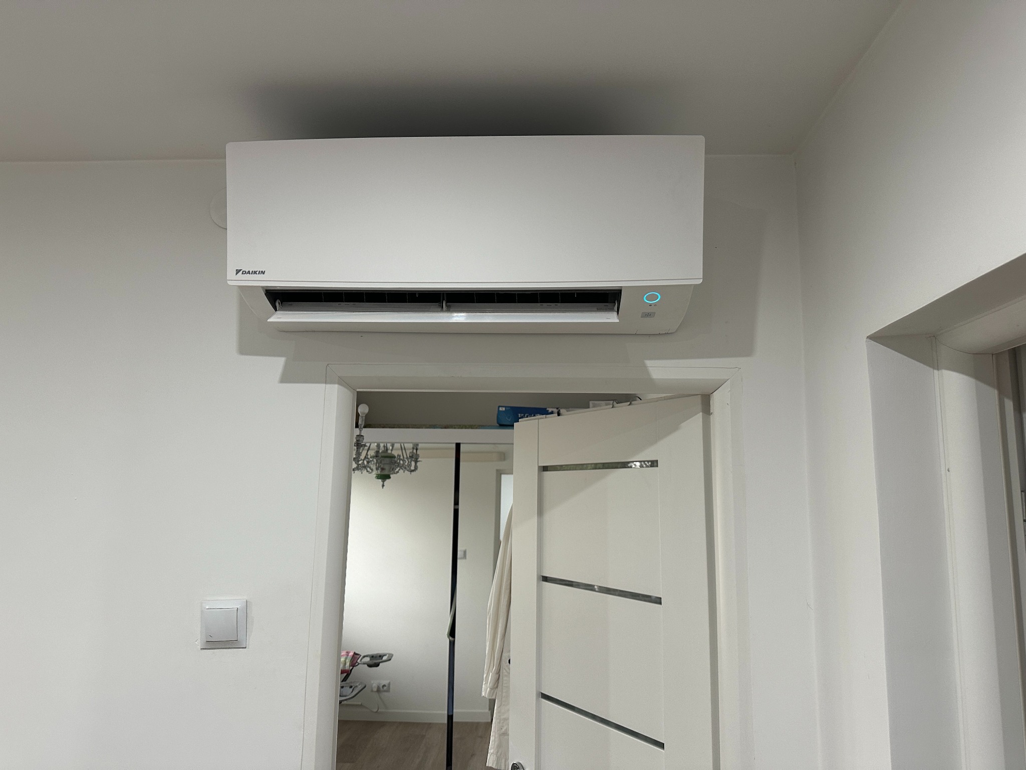 Biała klimatyzacja Daikin zamontowana na ścianie nad otwartymi drzwiami w jasnym wnętrzu. Widoczny włącznik światła i fragment żyrandola w sąsiednim pomieszczeniu.