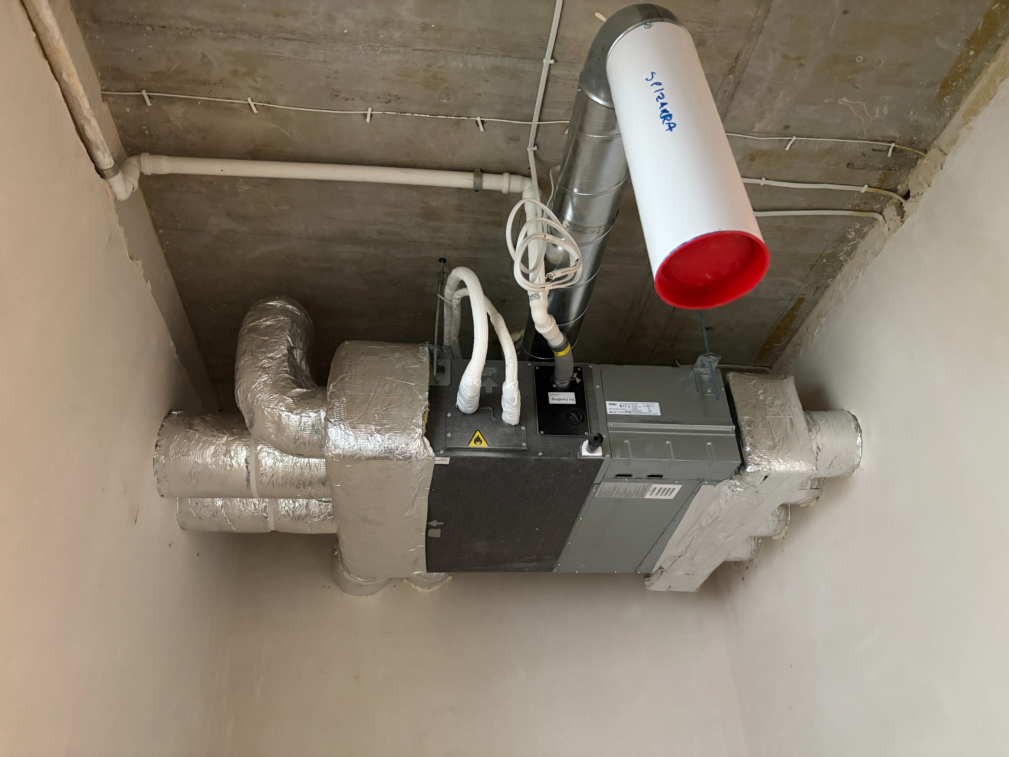 Centrala wentylacyjna zamontowana pod betonowym stropem, z widocznymi przewodami wentylacyjnymi owiniętymi folią izolacyjną oraz rurą z napisem 'SPIRARA'.