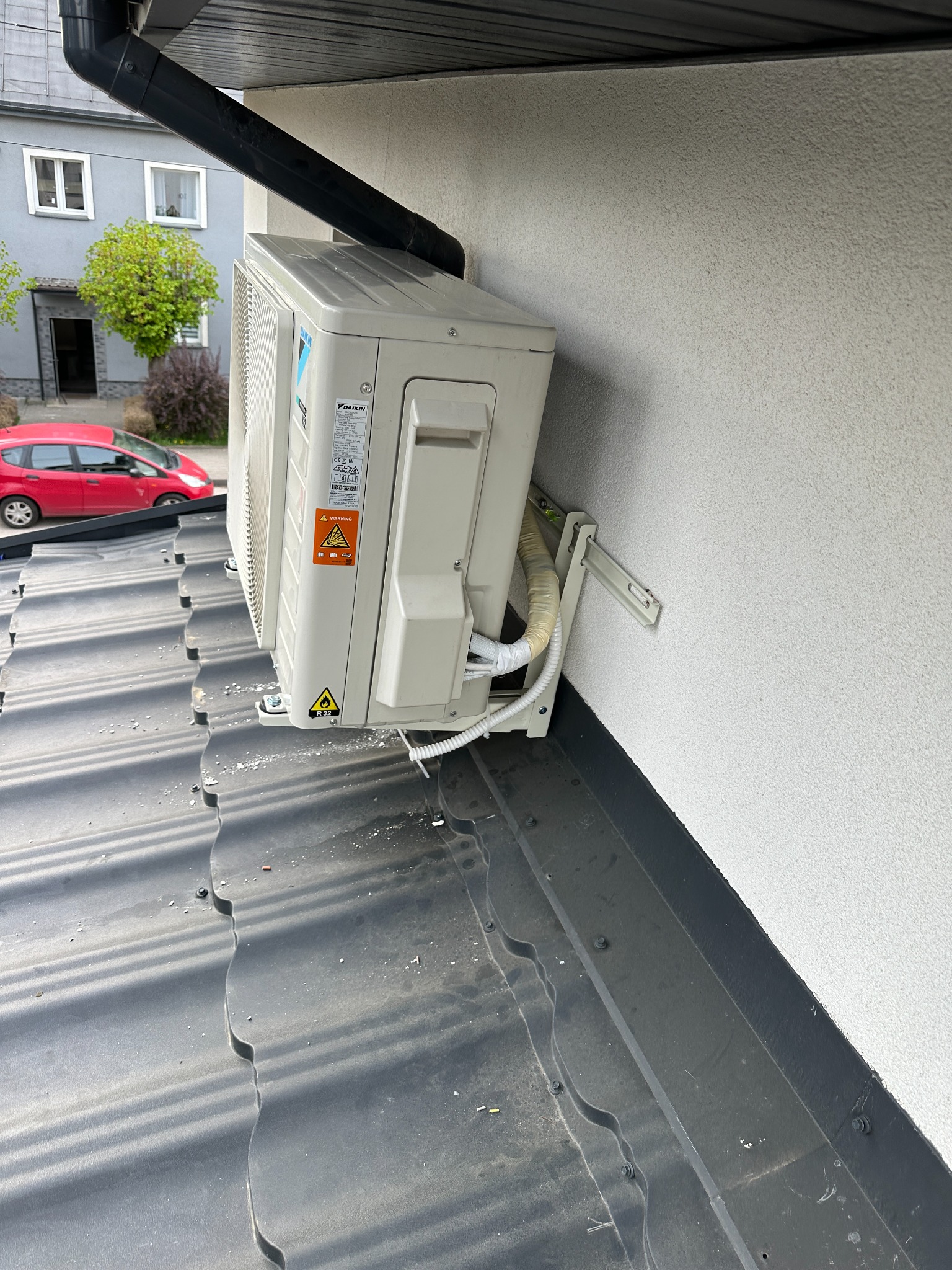 Zamontowana jednostka zewnętrzna klimatyzacji Daikin na dachu, widoczna instalacja i uchwyty montażowe. W tle budynek mieszkalny i czerwony samochód.