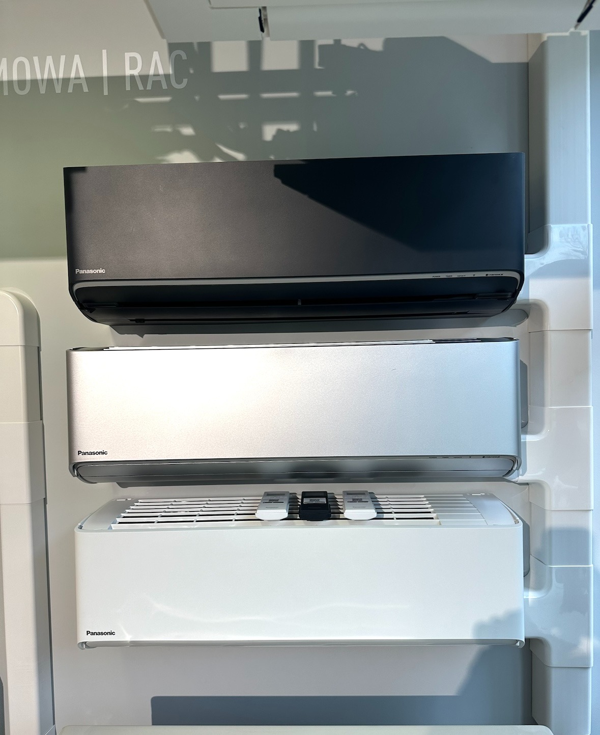Wystawa klimatyzatorów Panasonic w kolorach: czarnym, srebrnym i białym. Na białym klimatyzatorze leżą trzy piloty.