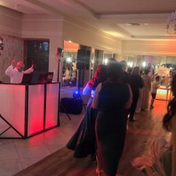 NEXT Party - DJ z podświetlanym stanowiskiem gra muzykę na parkiecie podczas imprezy, w tle bawią się goście. Widoczny sprzęt nagłośnieniowy i oświetleniowy.