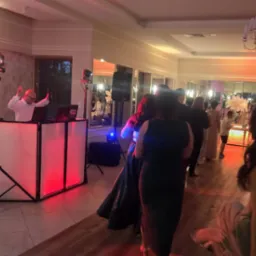 DJ z podświetlanym stanowiskiem gra muzykę na parkiecie podczas imprezy, w tle bawią się goście. Widoczny sprzęt nagłośnieniowy i oświetleniowy.